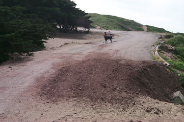 BEGINNING OF IMAGE DESCRIPTION.  IMAGE IS A PHOTOGRAPH OF THE ROAD HEADING DOWN TO THE JUNCTION OF TWO ROUTES AT FORT FUNSTON.  PHOTOGRAPH SHOWS A SECTION OF ROAD APPROXIMATELY THE AREA OF A CITY BUS THAT HAS ERODED.  HOWEVER, THIS HAS BEEN FILLED IN WITH DIRT AND GRAVEL SO THAT THE ROAD IS ALMOST AS WIDE AS IT IS IN THE NON ERODED SECTION.  END OF IMAGE DESCRIPTION.